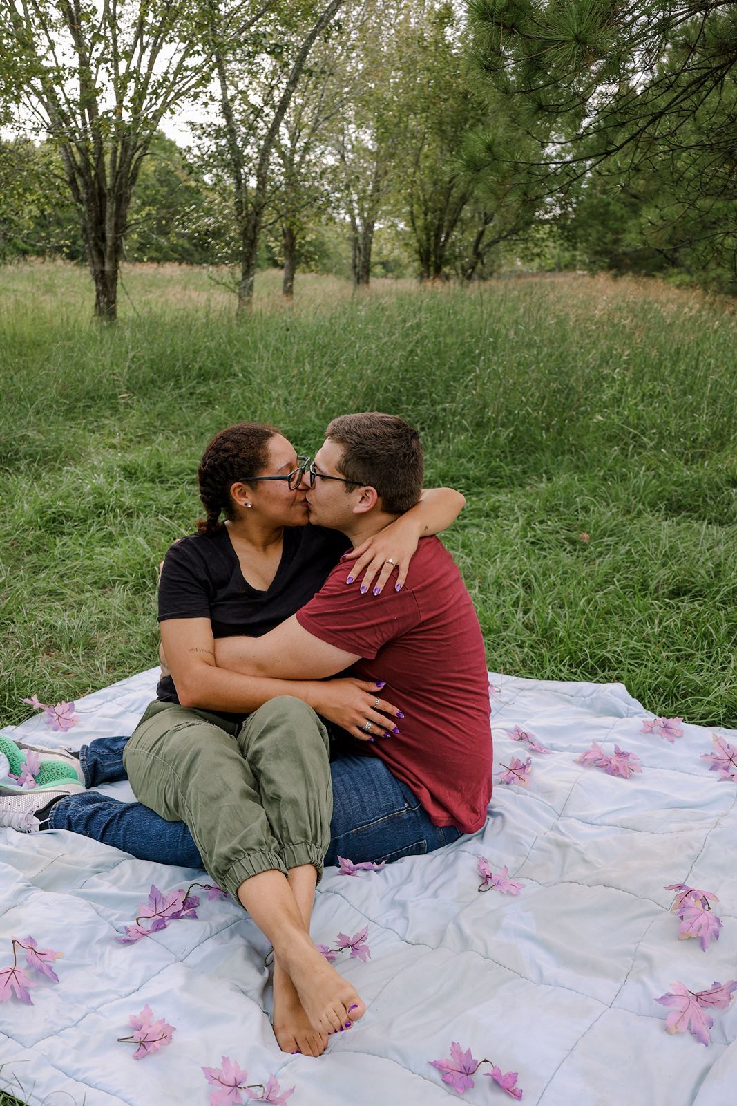A happy couple shares a kiss while sitting on a floral blanket in a lush green outdoor setting.