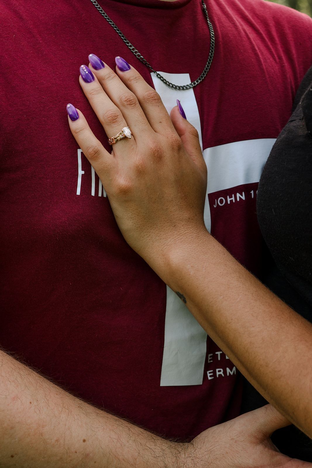 Close-up of a woman's hand with purple nails and a gold ring resting on a man's red graphic t-shirt.