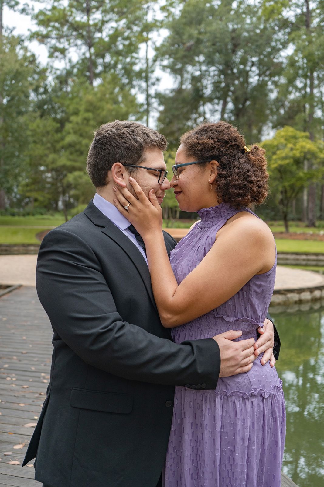 Romantic couple nearly kissing on a park dock, man in dark suit, woman in purple sleeveless dress.