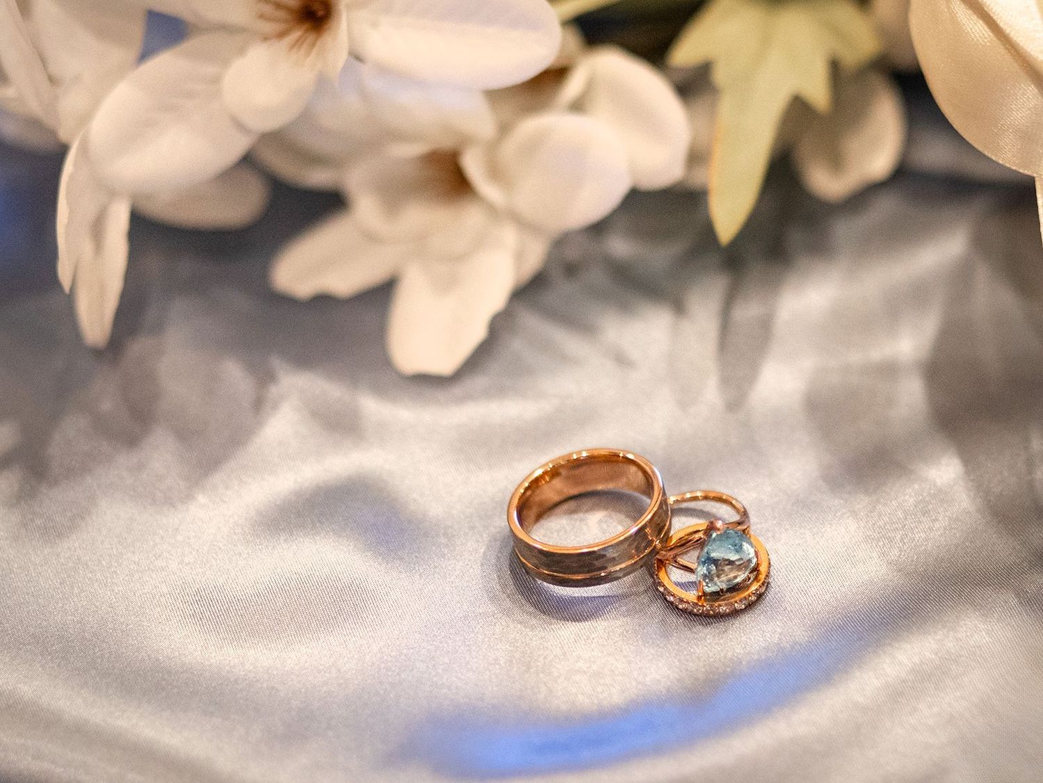 Two gold wedding rings resting on silver satin fabric with white flowers in the background.