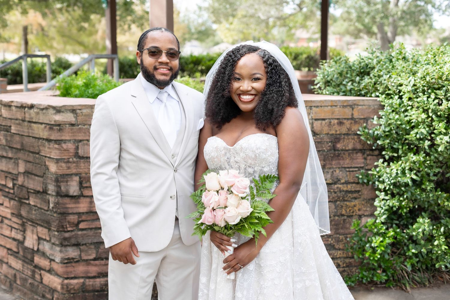 Elegant wedding couple poses outdoors against lush greenery and stone walls in summery garden setting.