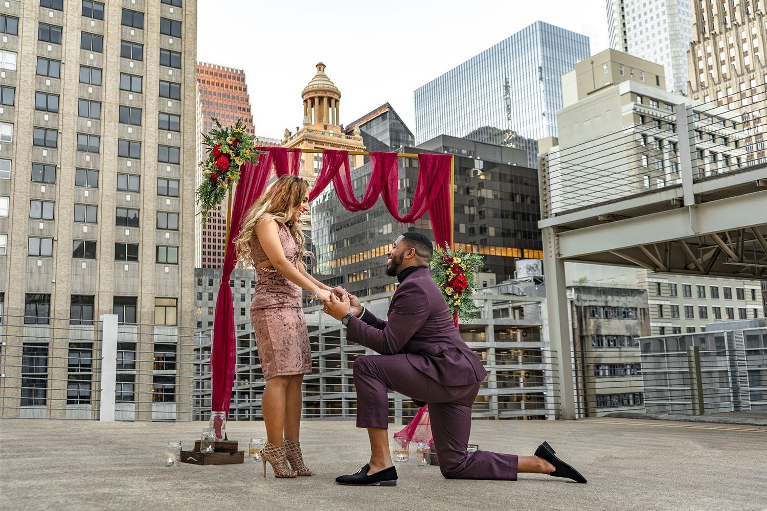 A romantic rooftop proposal scene with a decorated red and gold mandap against a city skyline backdrop.
