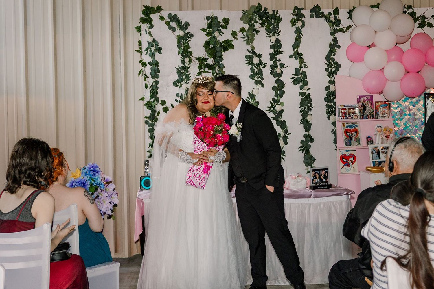 Couple shares a kiss at quinceañera celebration with floral backdrop, balloons, and seated guests.