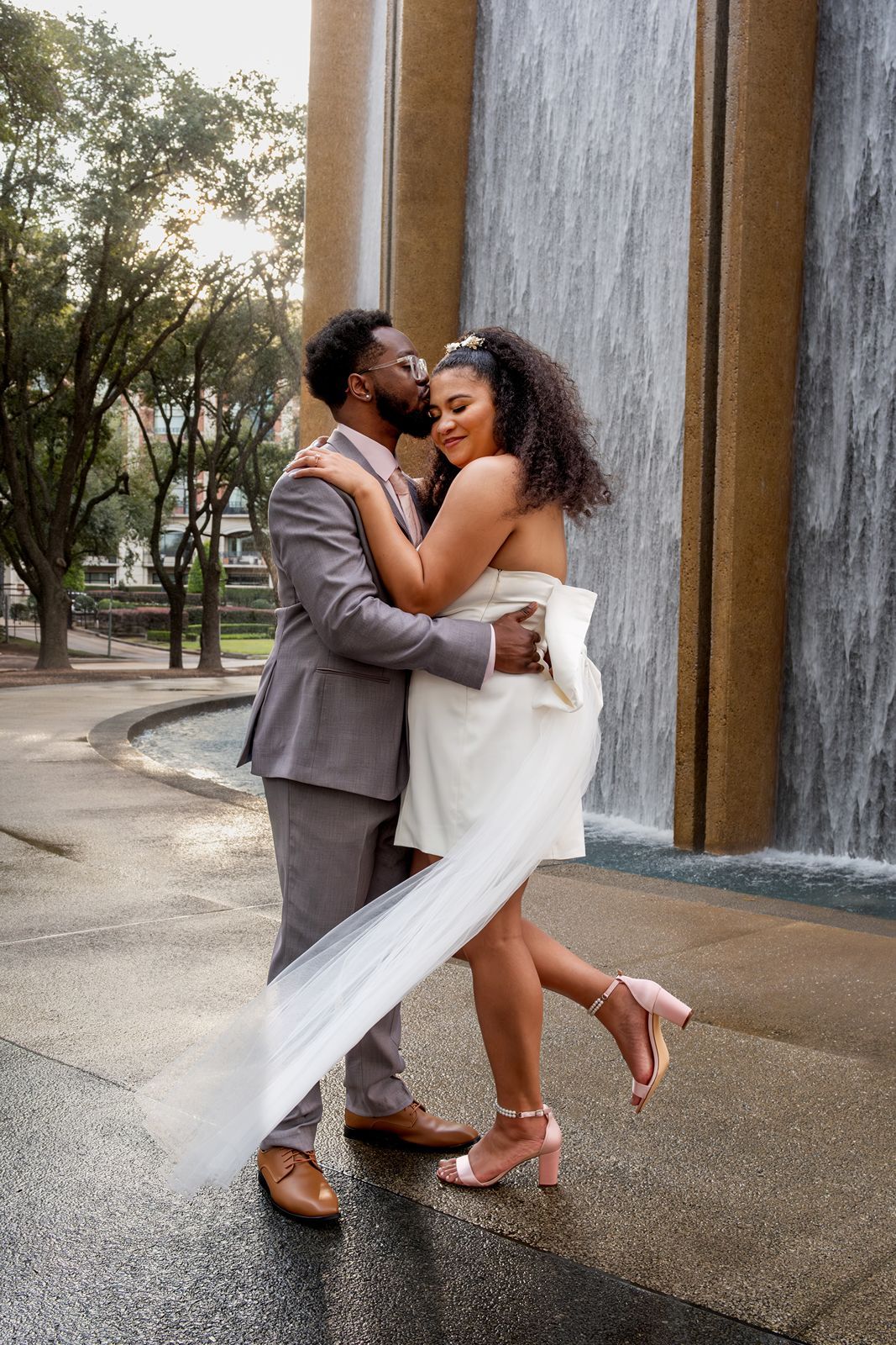 A romantic couple embraces and kisses in front of a waterfall wall at an outdoor venue.