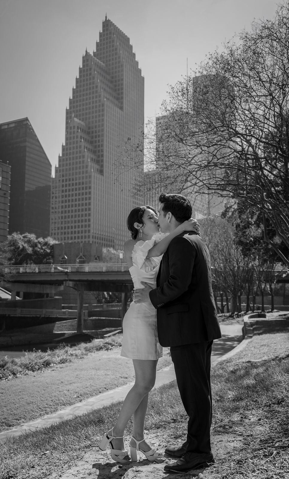 A couple shares a romantic moment in black and white against the backdrop of modern skyscrapers in a city park.
