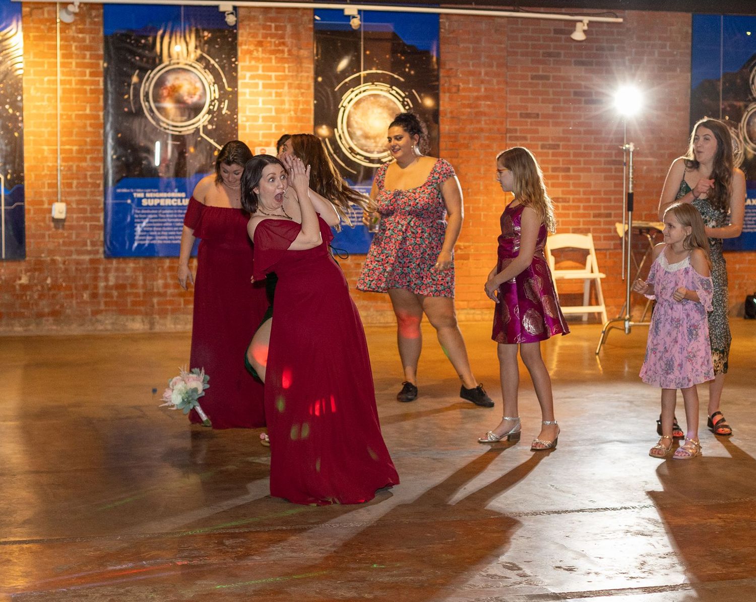 Bridesmaids in red dresses dance joyfully at a wedding reception in a rustic brick venue with warm lighting.