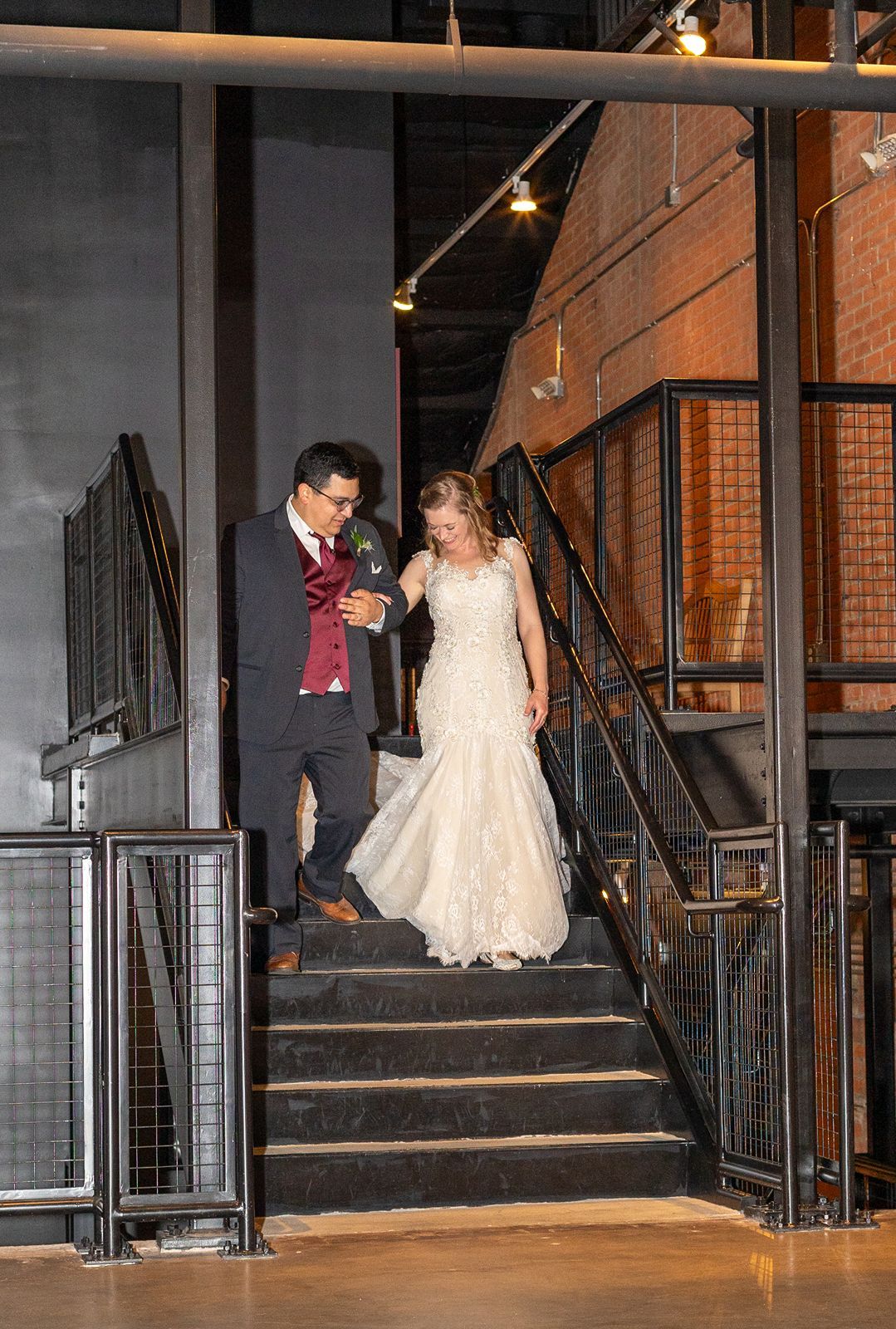 Bride in lace mermaid gown and groom in burgundy vest descend industrial staircase at wedding reception.