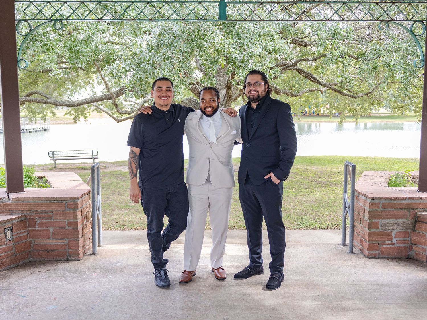 A wedding party takes casual photos under a covered outdoor pavilion with brick pillars.