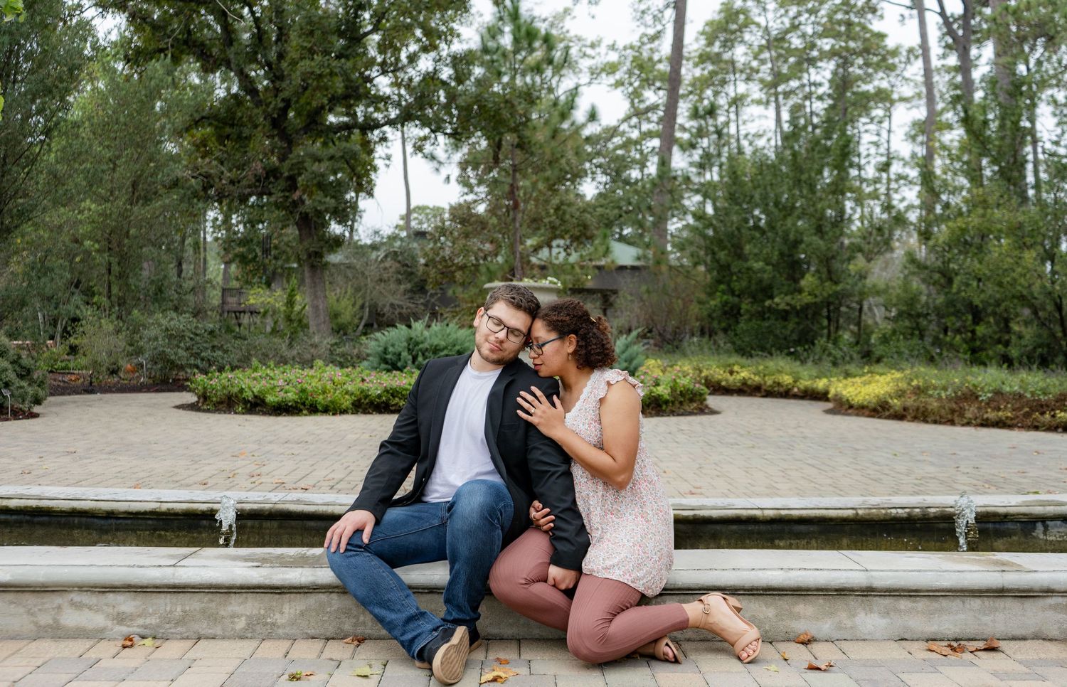 A romantic engagement photo on a park bench surrounded by pine trees.
