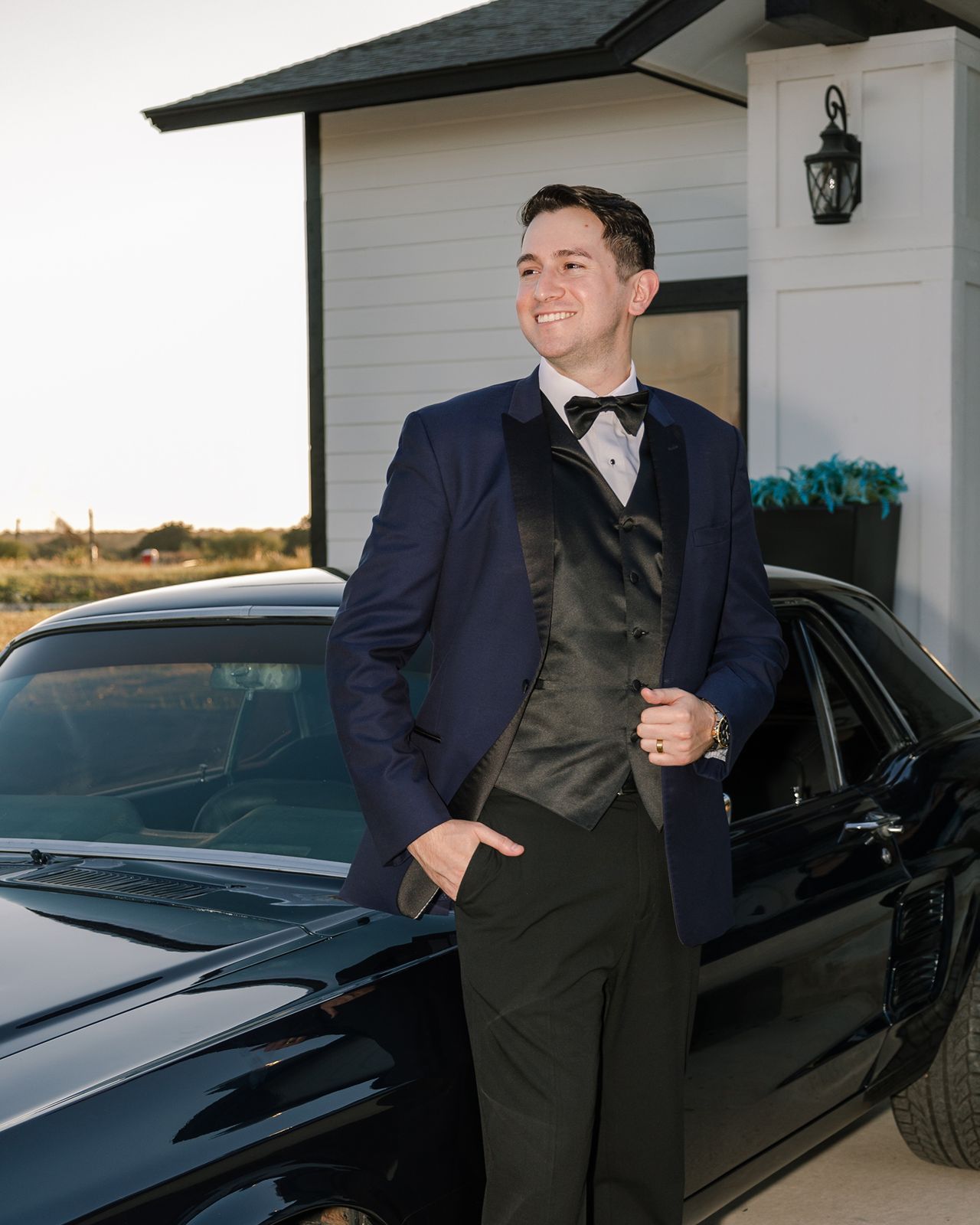 A groom in a navy suit poses playfully by a black luxury car during a wedding photoshoot.