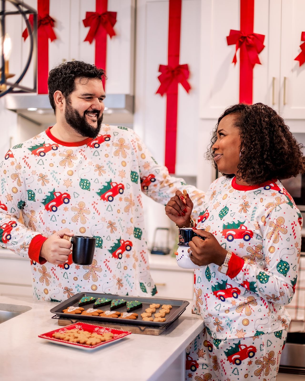 People wearing matching holiday pajamas with red truck patterns share a joyful moment in the kitchen.