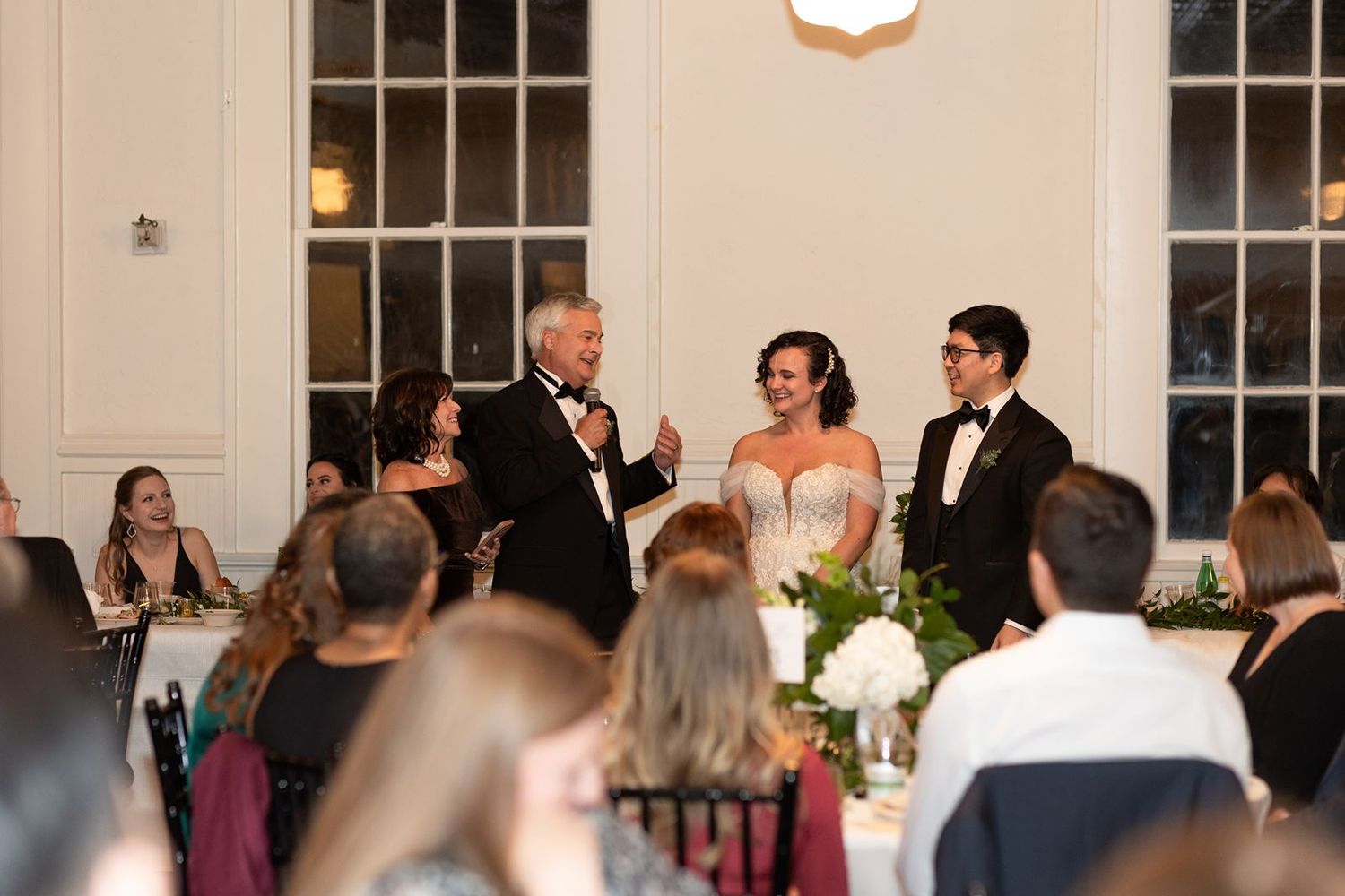 Wedding reception guests seated at round tables in an elegant ballroom with large windows and chandeliers.