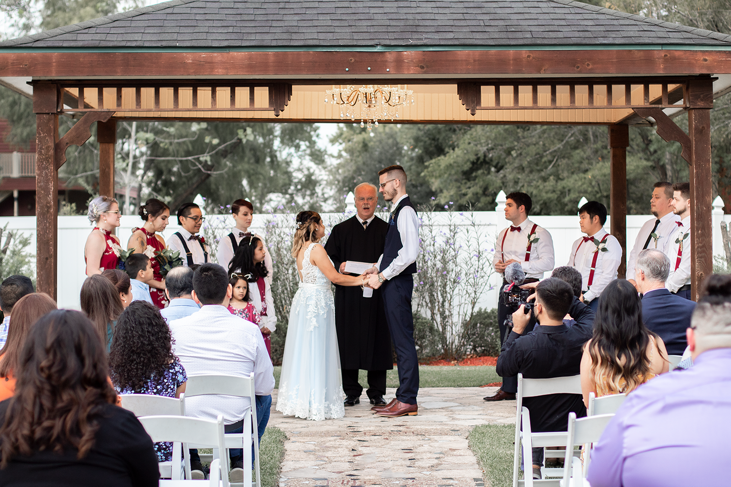 Outdoor wedding ceremony under a wooden gazebo with bride, groom, officiant, and wedding party on a sunny day.