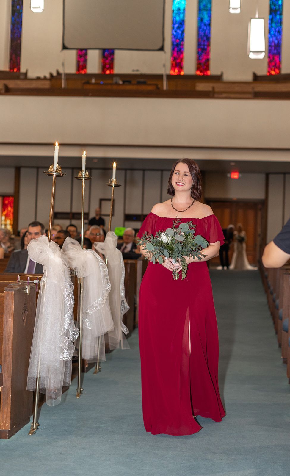 Bridesmaid in red off-shoulder gown walks down church aisle holding bouquet during wedding ceremony.