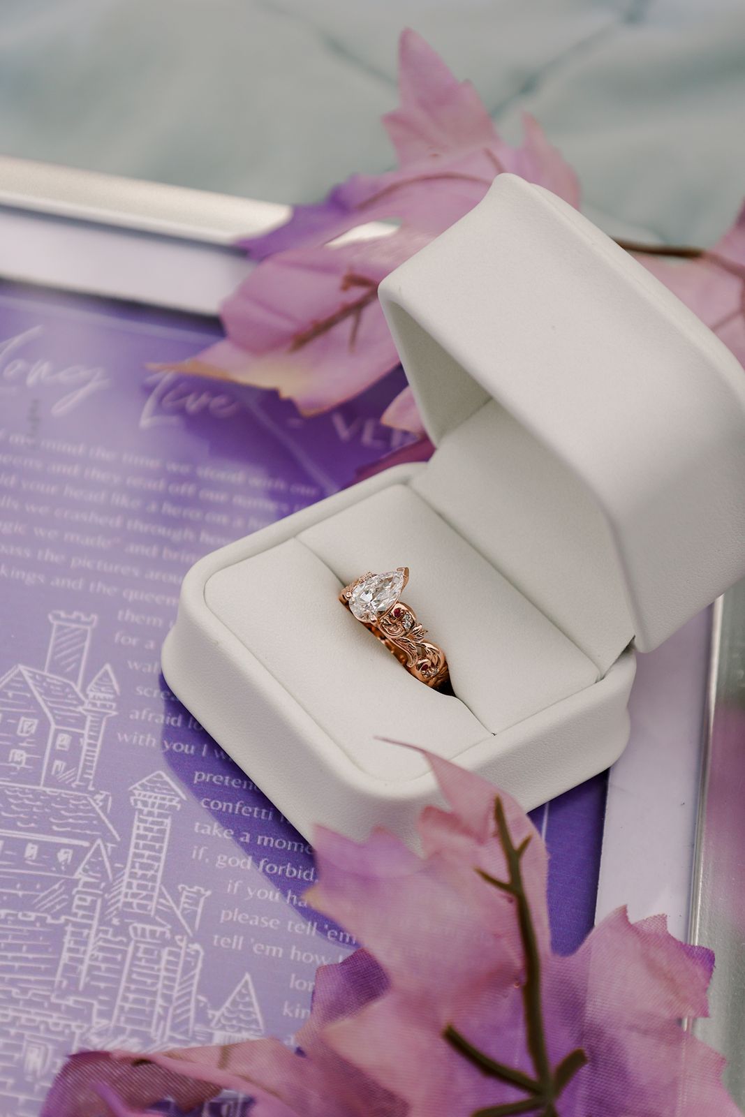 A rose gold diamond engagement ring in a white velvet box surrounded by purple flowers and a love letter.