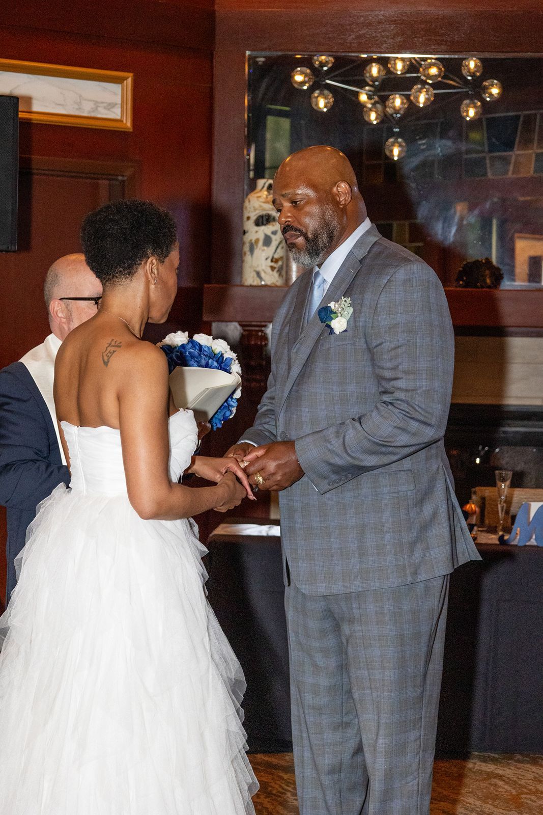 Groom in gray plaid suit places wedding ring on brides finger during indoor ceremony.