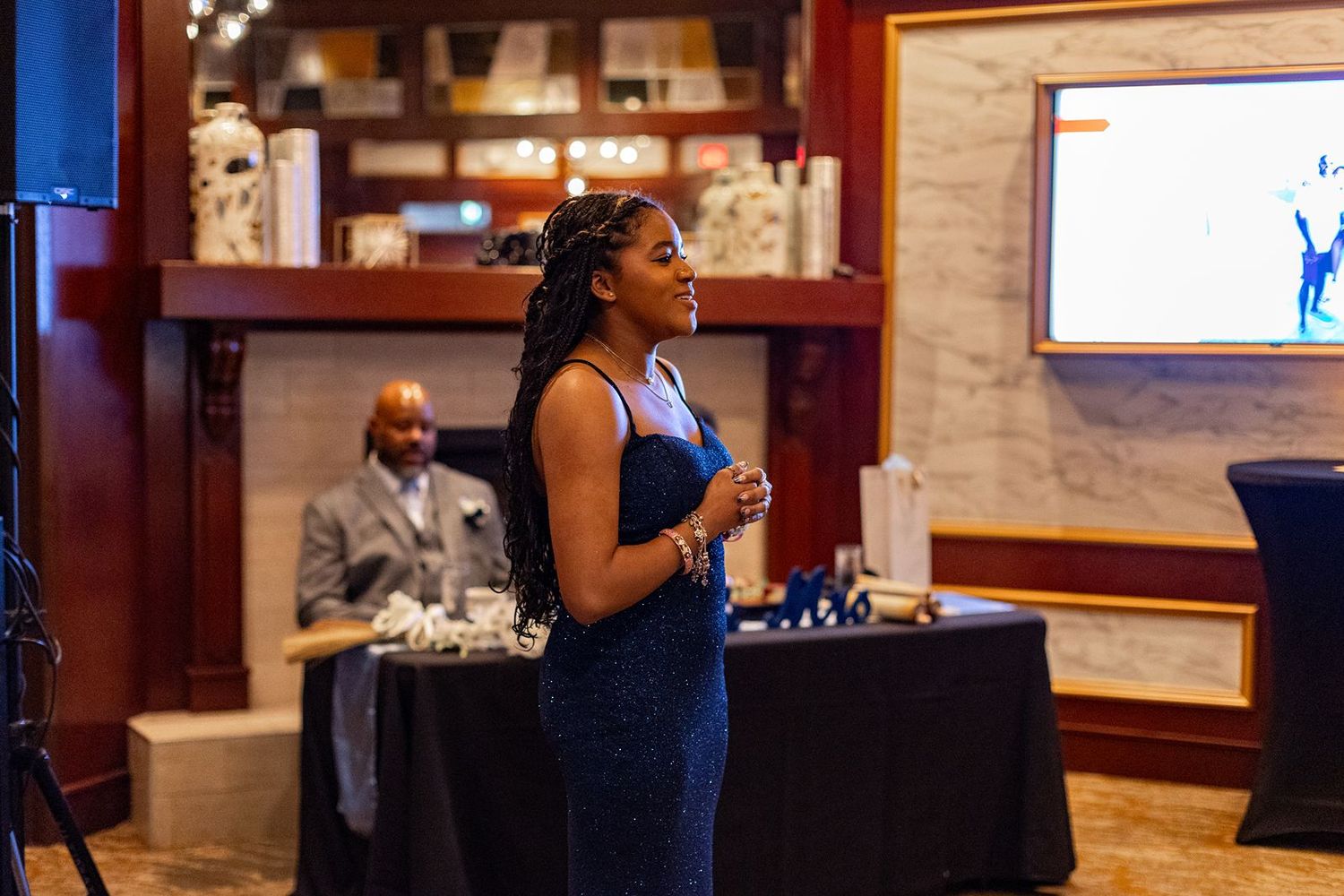 Woman in navy blue dress speaks at an upscale event venue with guests seated in background.