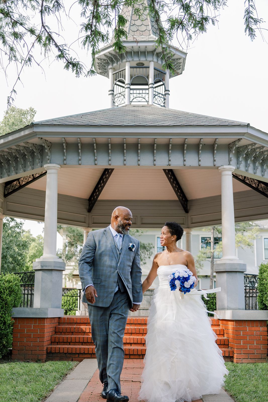 Newlywed couple walking hand in hand down brick steps from a white Victorian gazebo on their wedding day.