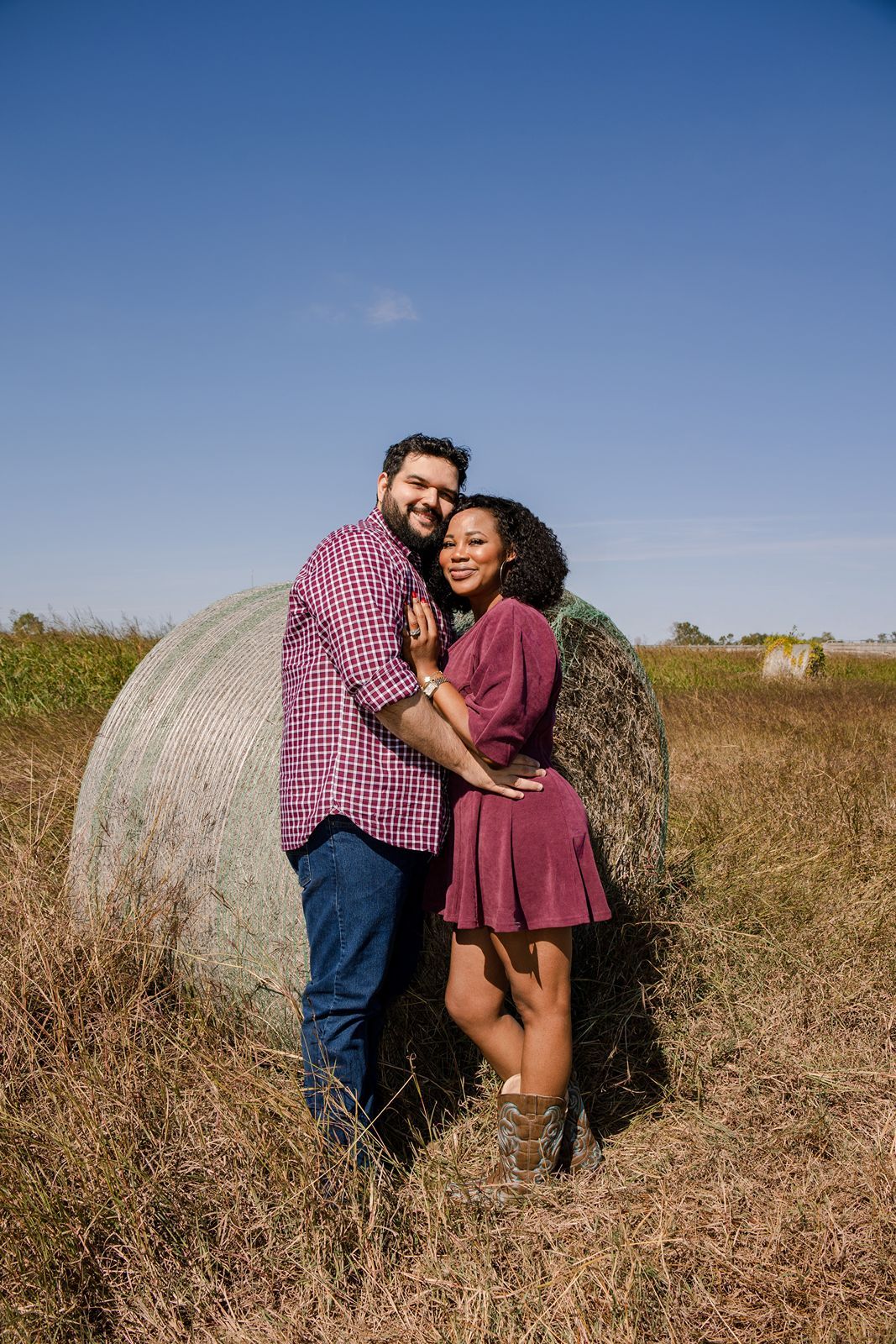 A couple embraces and laughs together in a rustic field setting with a large hay bale in the background.