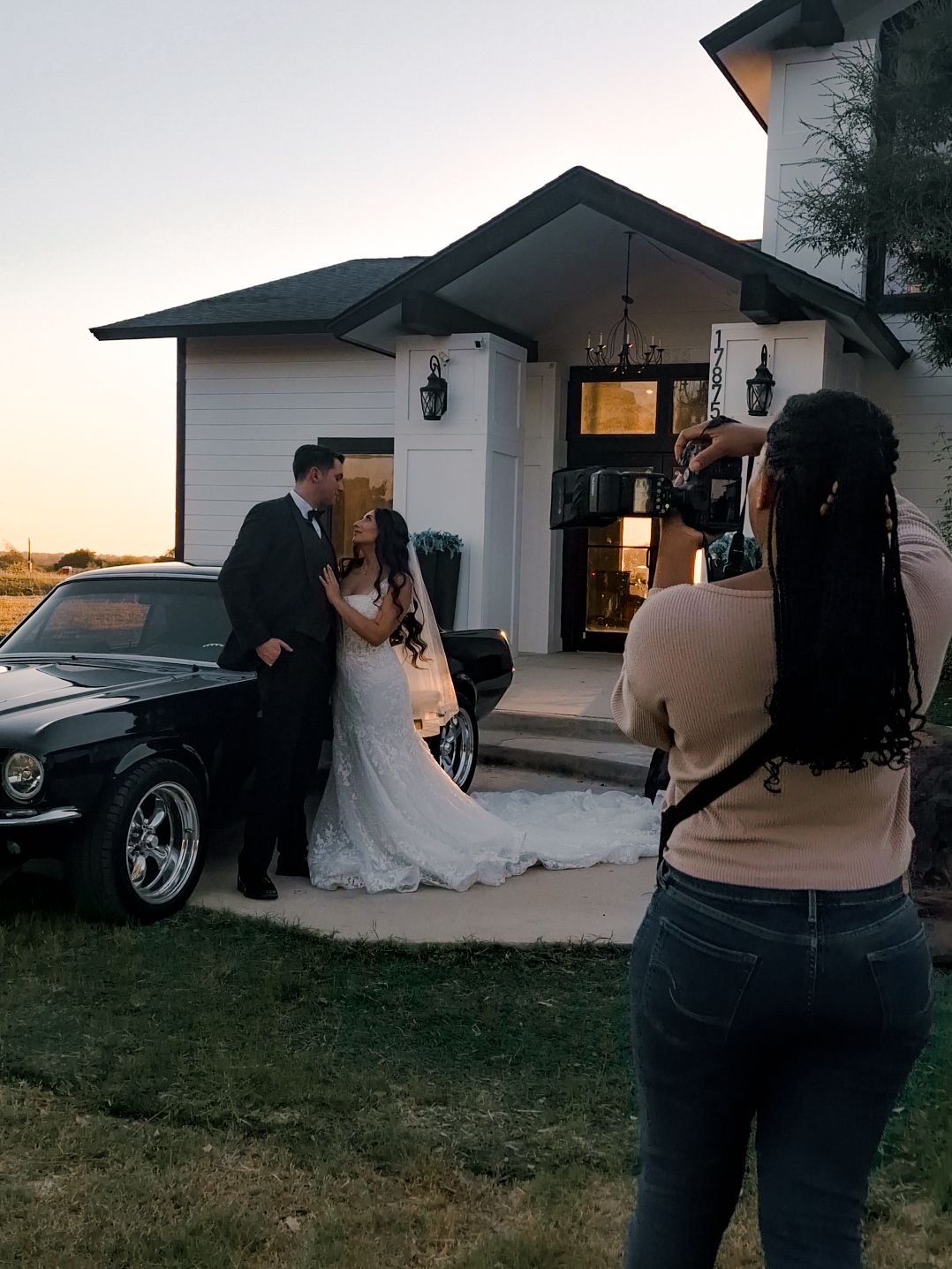 Photographer capturing wedding couple in formal attire posing by classic black car at sunset outside white house.