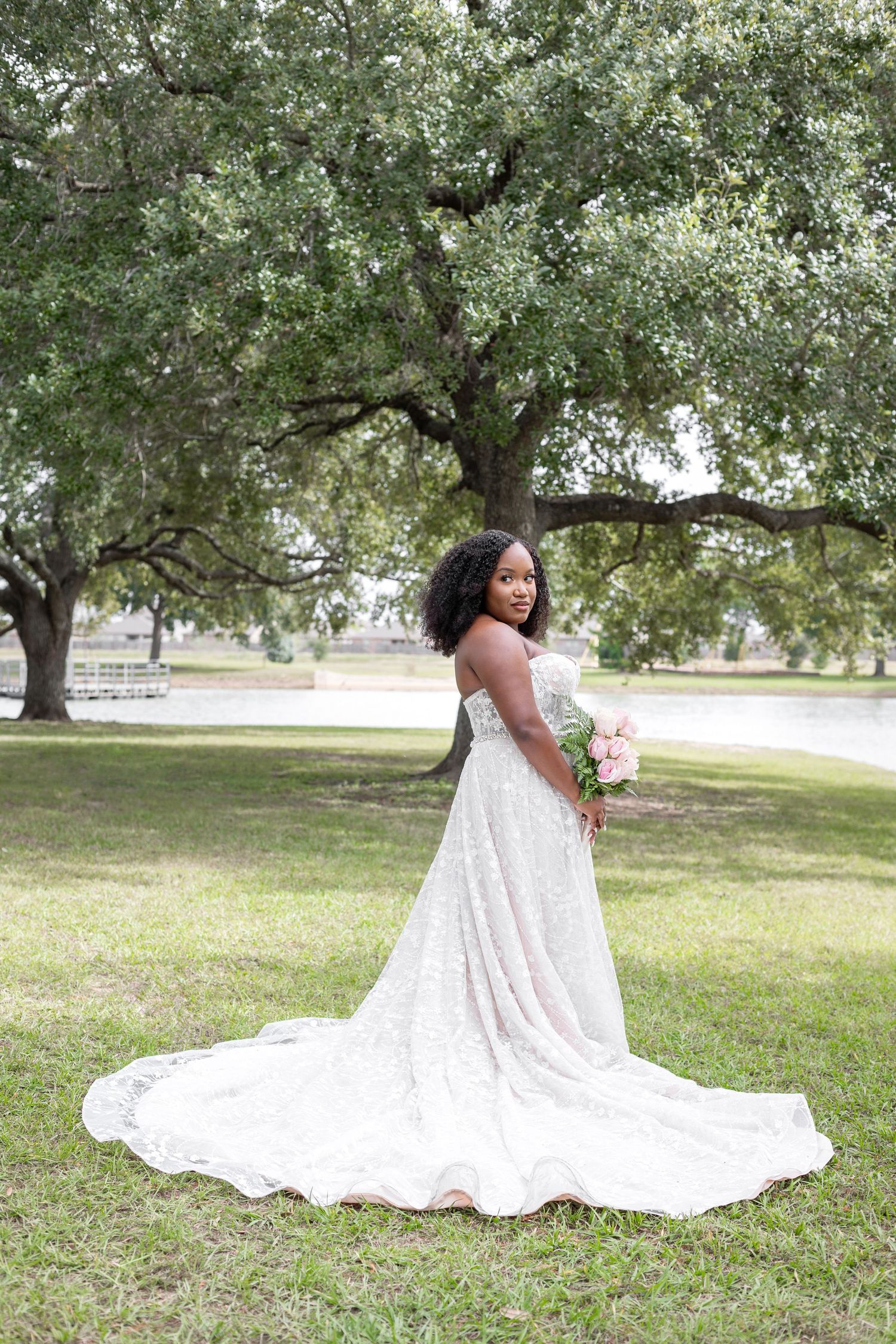 Beautiful wedding dress with flowing train spreads across green grass under oak trees in outdoor photo session.