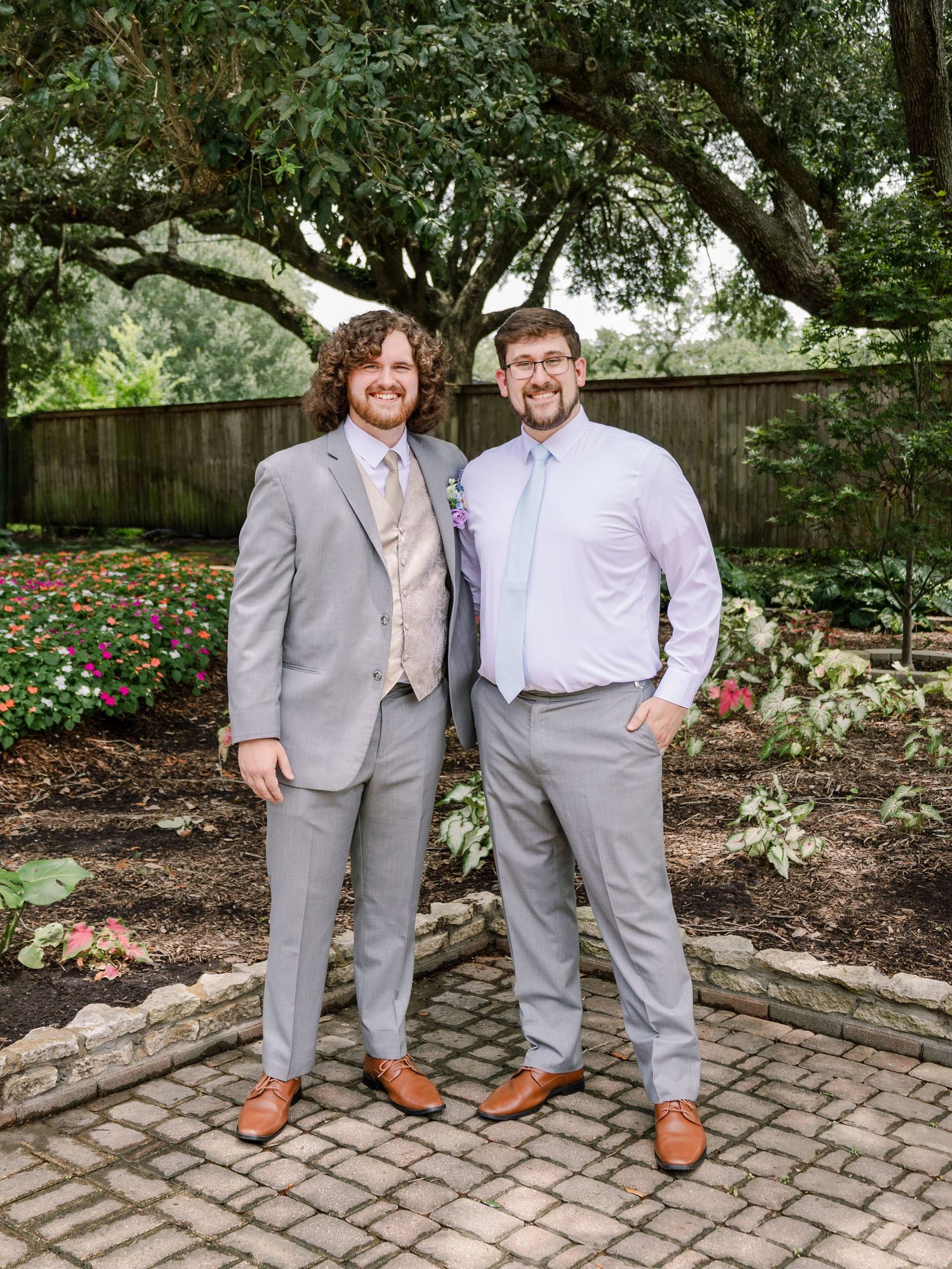 Groomsmen in gray suits and pink ties posing together on brick pavers in a garden.