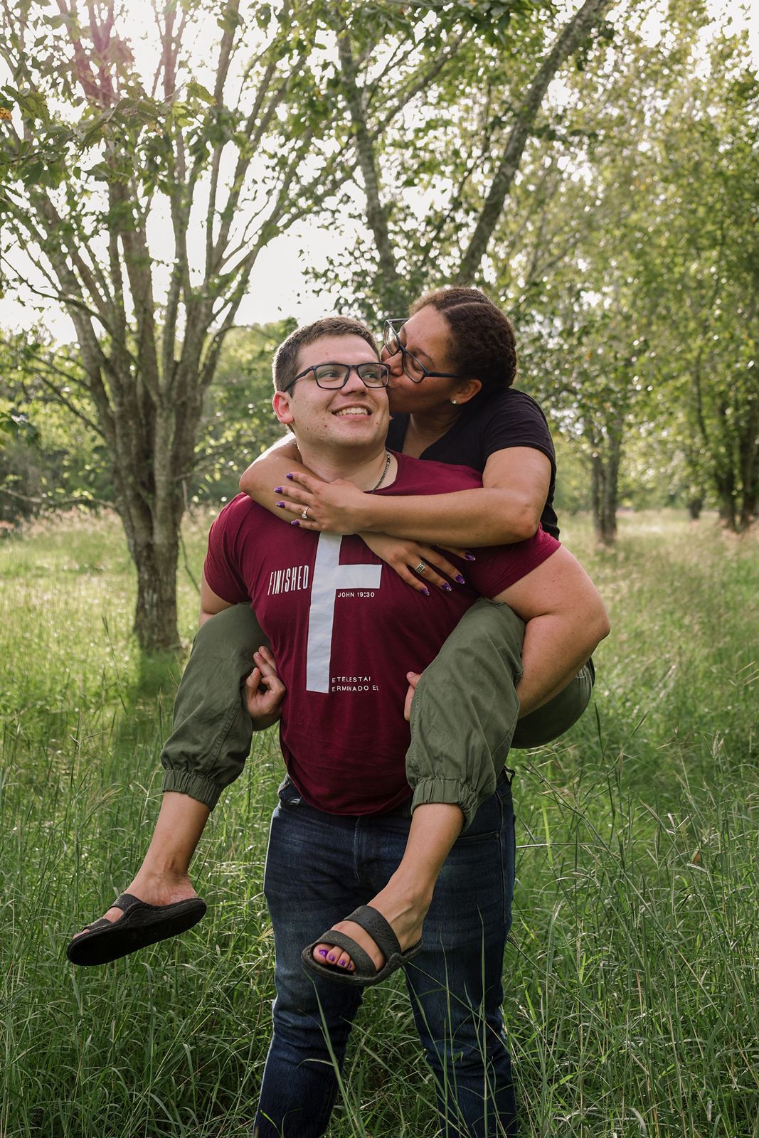 A couple shares a joyful piggyback ride in a lush green park, the woman kissing the mans cheek as he smiles.