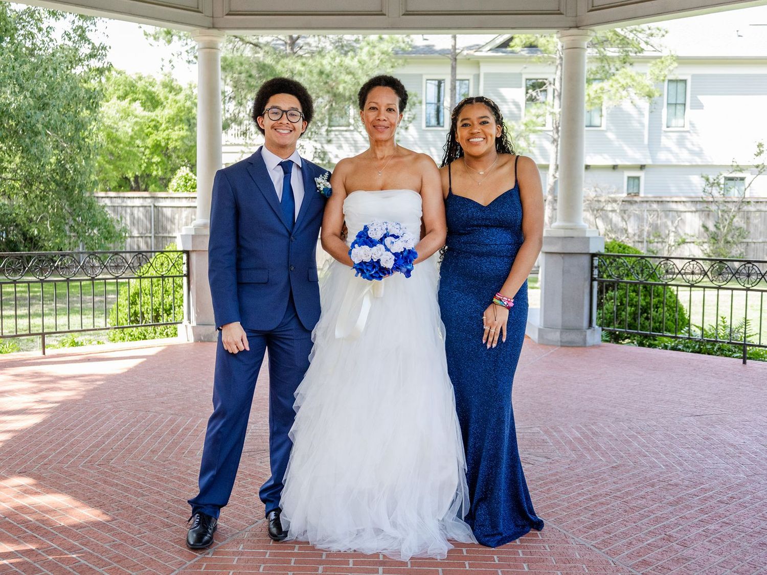 Bride in white gown poses with two guests in blue attire under gazebo.