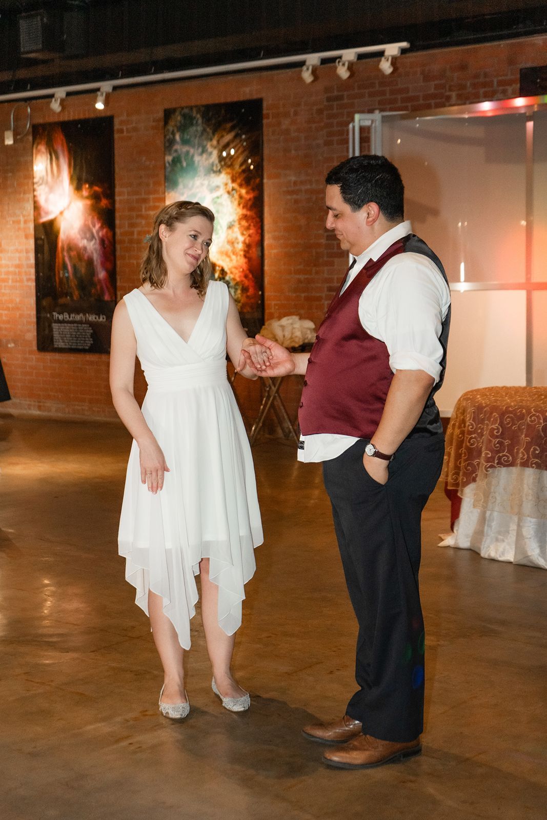 Bride in white high-low dress and groom in burgundy vest share first dance in rustic brick venue.