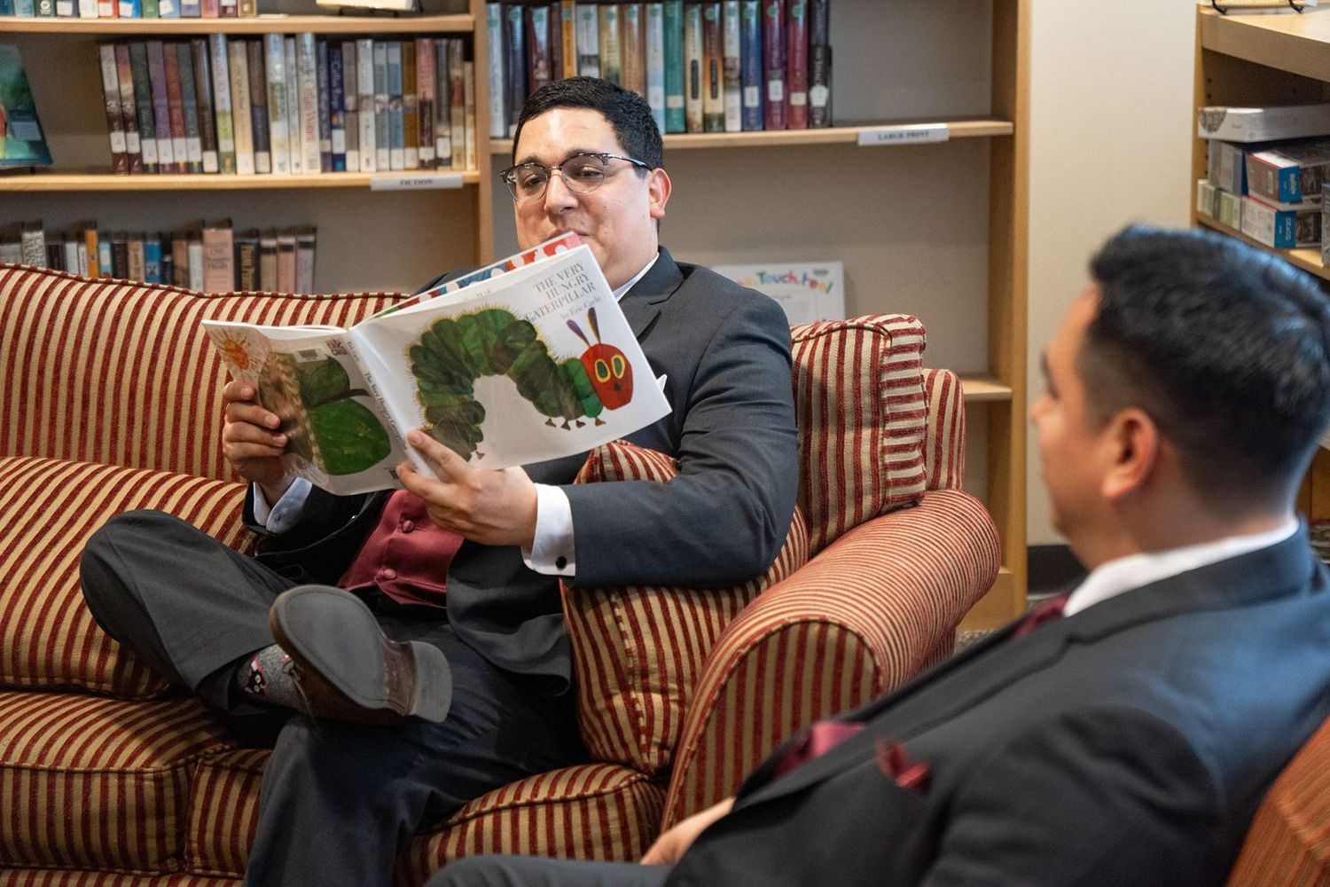 Two men in suits sit in a library, one reading The Very Hungry Caterpillar book, smiling in a cozy setting.