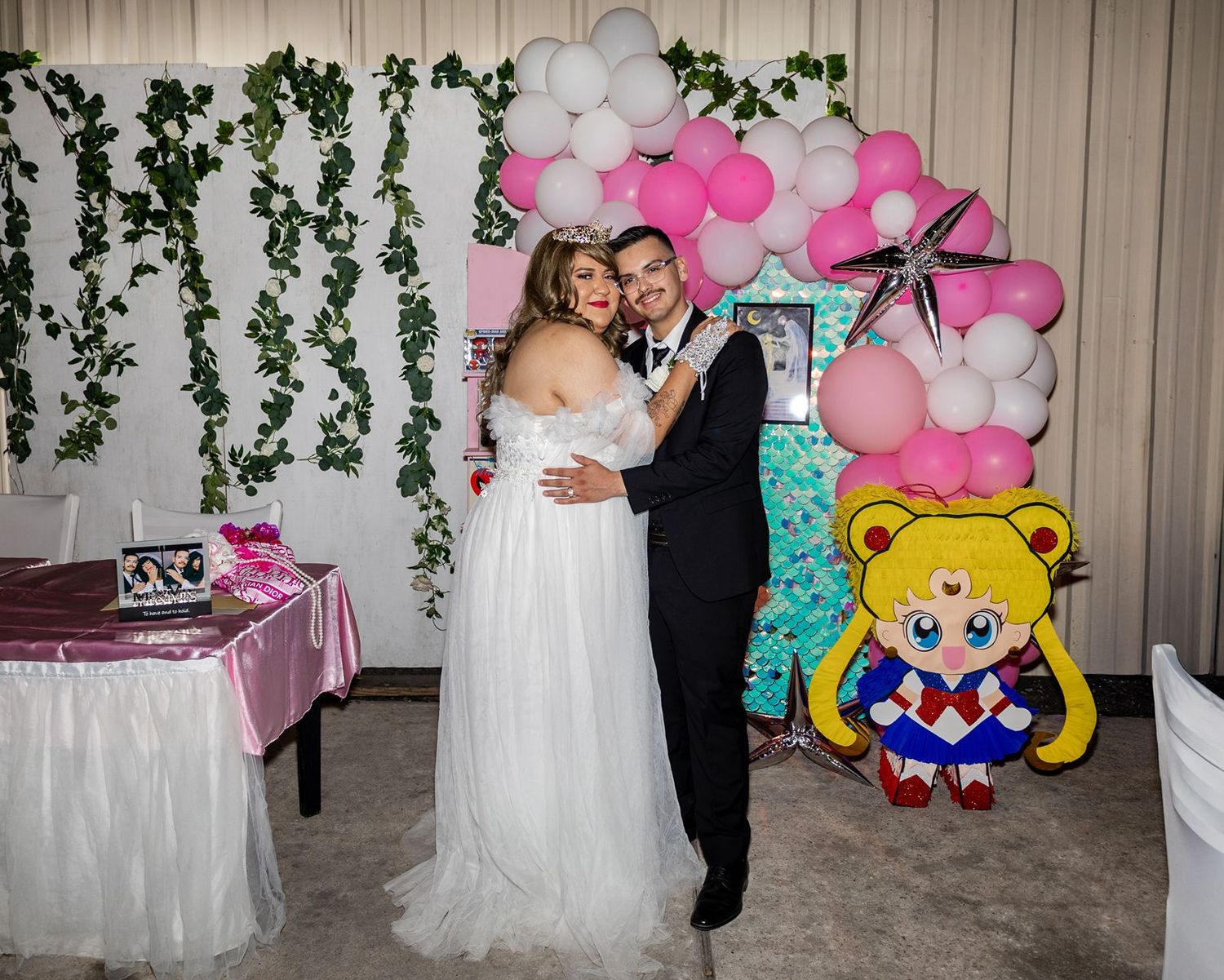 Couple poses in wedding attire before pink balloon arch with Sailor Moon decoration.