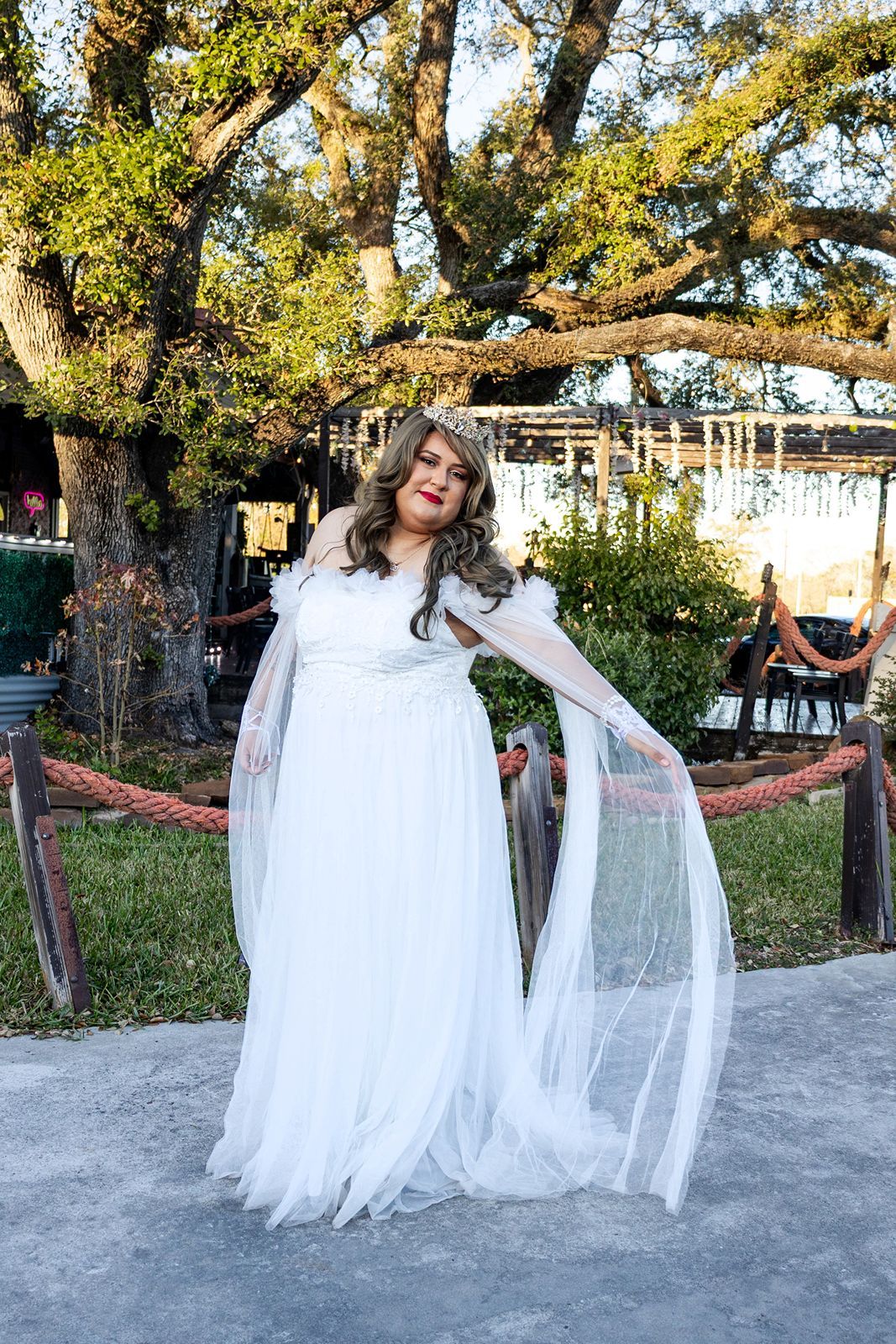 Woman in elegant white off-shoulder bridal gown with flowing tulle cape poses outdoors near large oak tree.