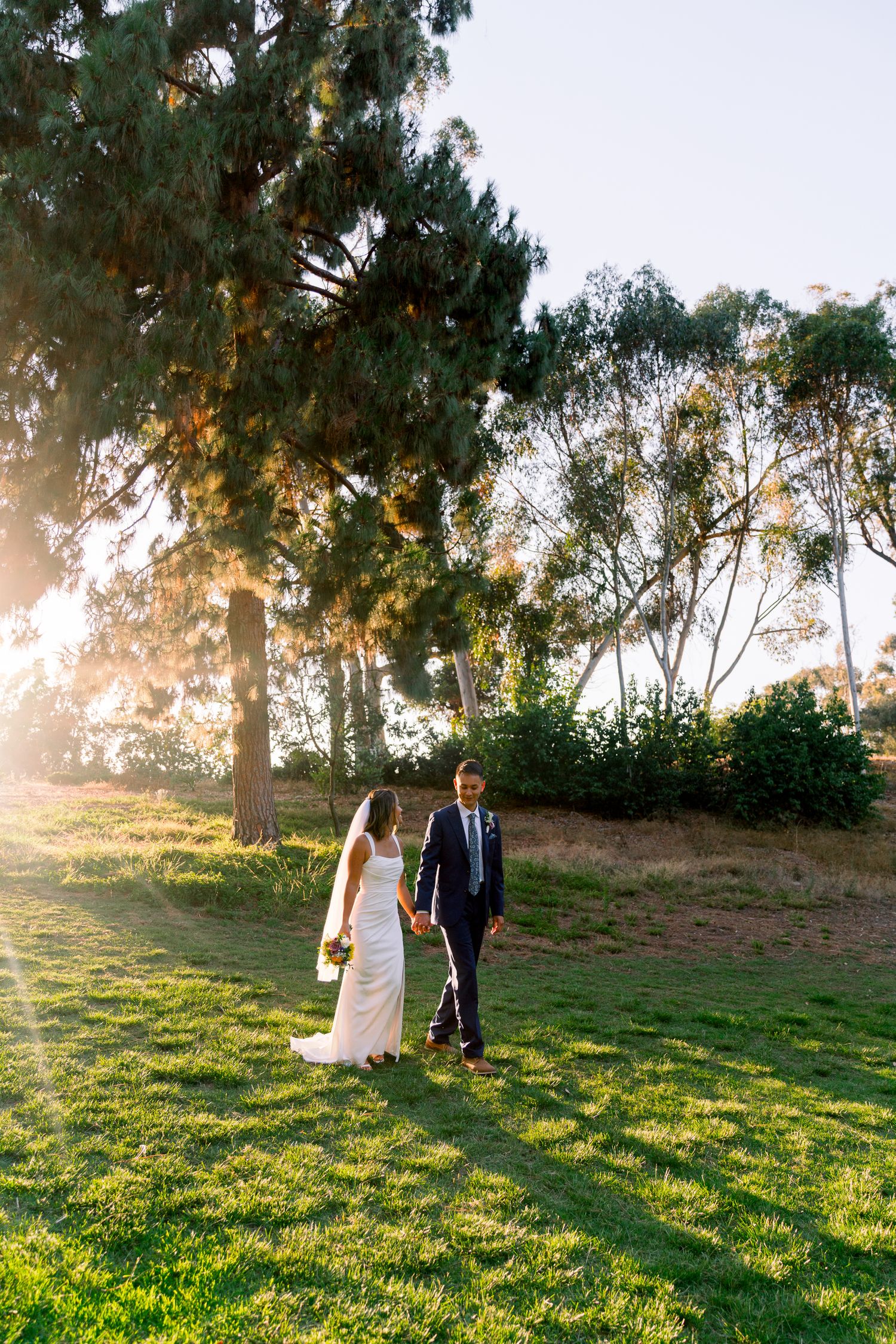 Two figures in wedding attire walking on a sunlit grassy lawn with tall evergreen trees in the background at sunset.