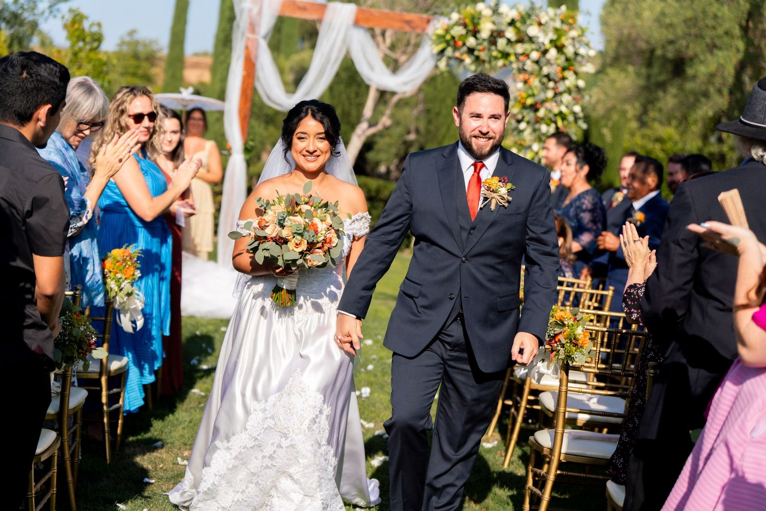 A happy couple walking down the aisle while wedding guests cheer during an outdoor ceremony.