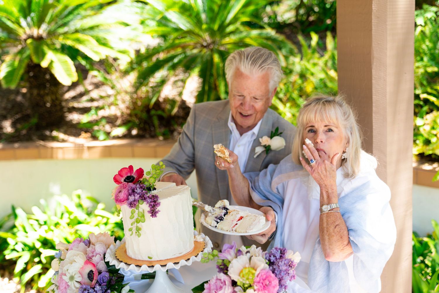 A happy couple shares a joyful moment while cutting their wedding cake, surrounded by purple floral decorations.