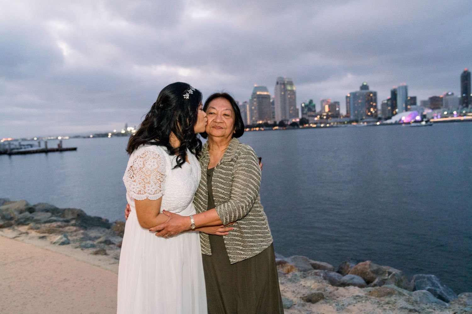 Two people embrace on a rocky shore with a city skyline across the water on a cloudy day.