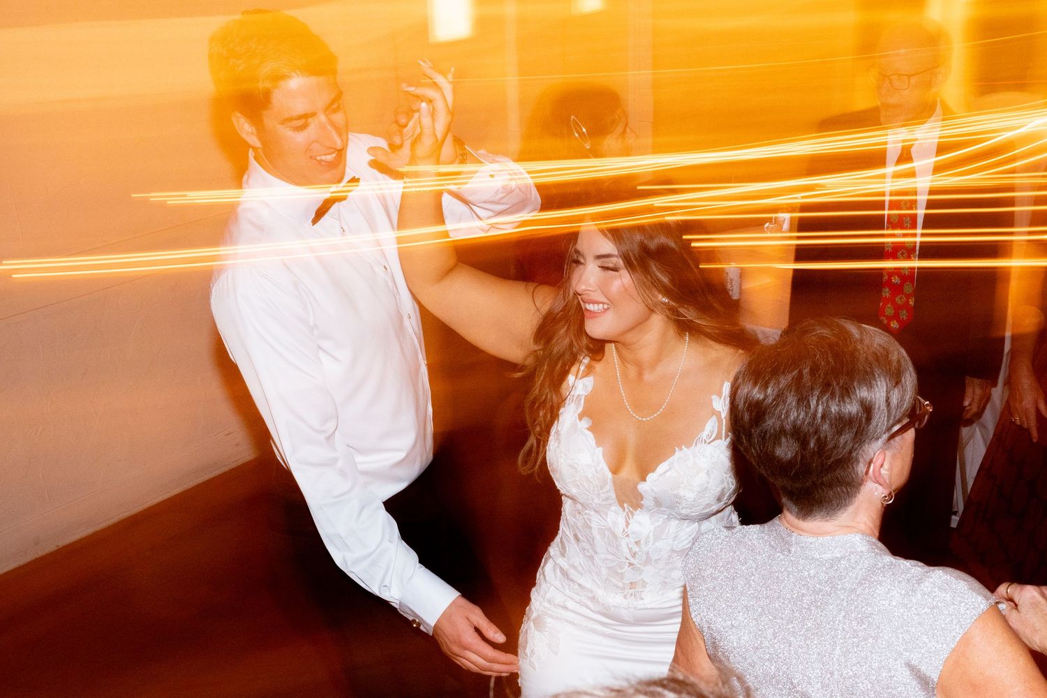 Wedding guests dancing energetically at a reception with motion blur and warm orange lighting.