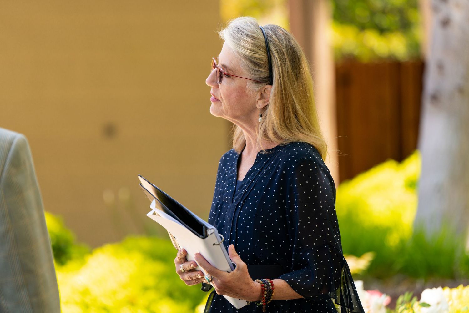 A woman in a polka dot blouse holds a tablet while standing outside on a sunny day.