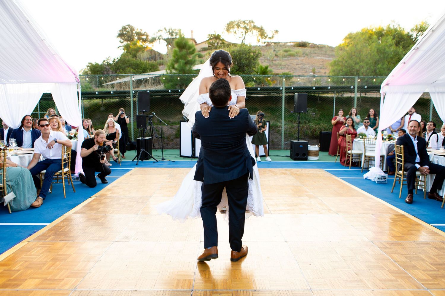 Two figures share an intimate slow dance together on a wooden dance floor under an elegant reception tent at an outdoor event.