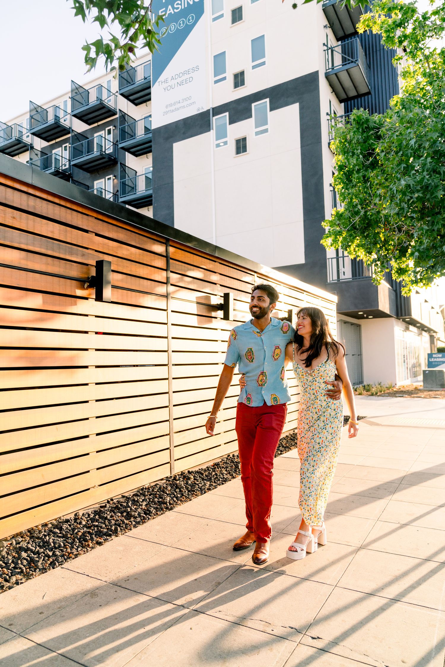 Couple walking hand in hand past modern apartment building with wooden fence.