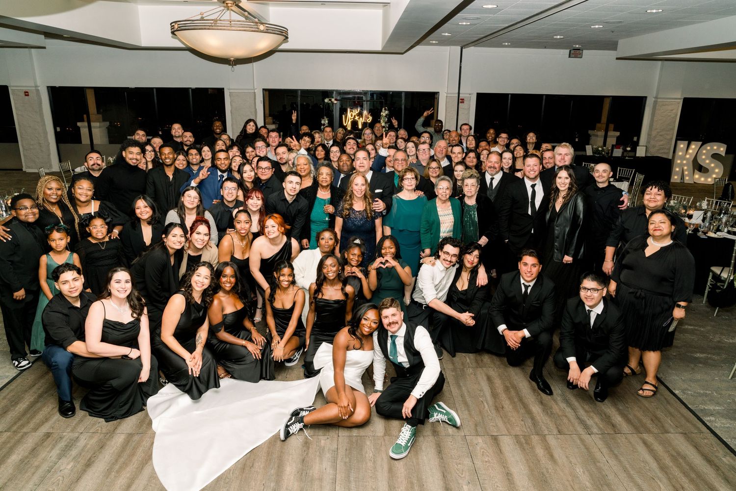 Large group wedding photo with guests in formal attire on dance floor.