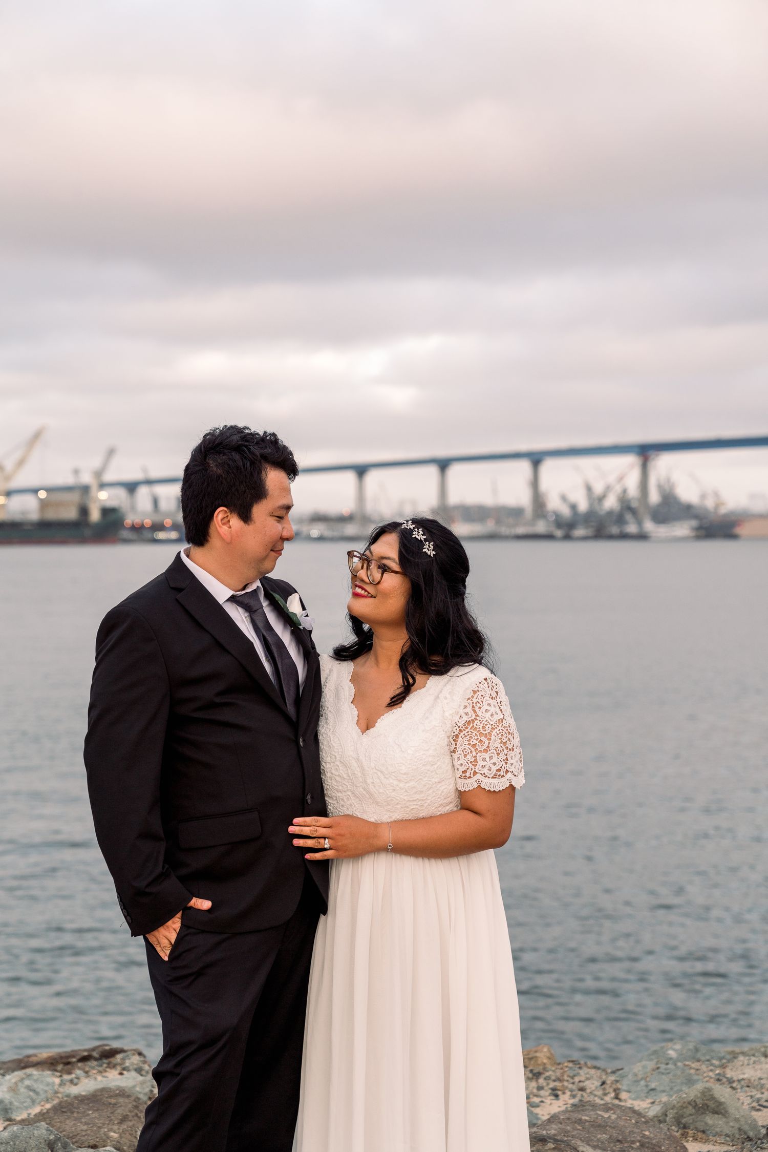 A romantic wedding couple shares an intimate moment by the waterfront with a bridge and industrial port in the background.