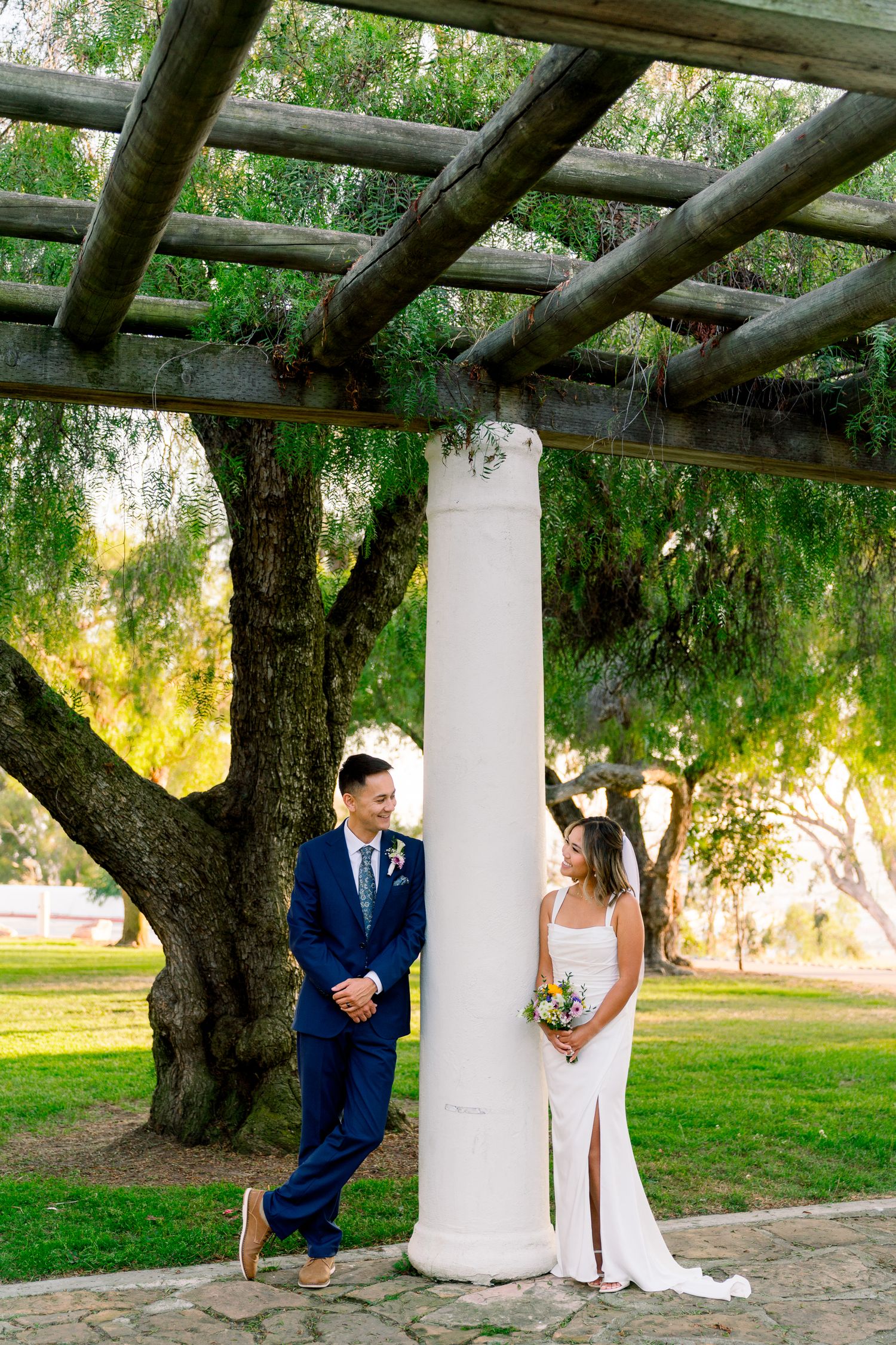 A couple poses beneath a rustic wooden pergola with white columns in a garden setting during sunset.