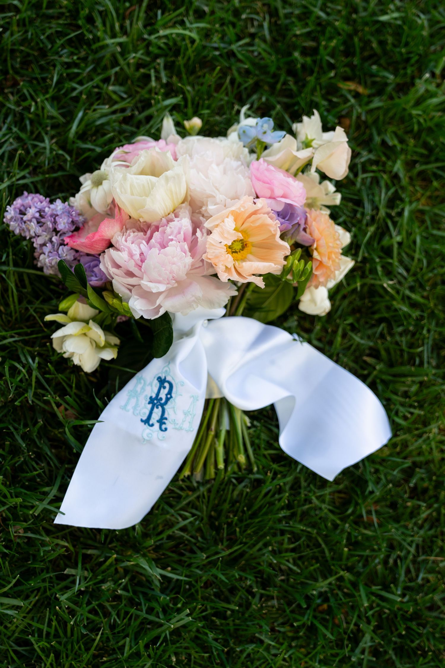A bridal bouquet of pink, peach and purple flowers with a monogrammed white ribbon lying on grass.