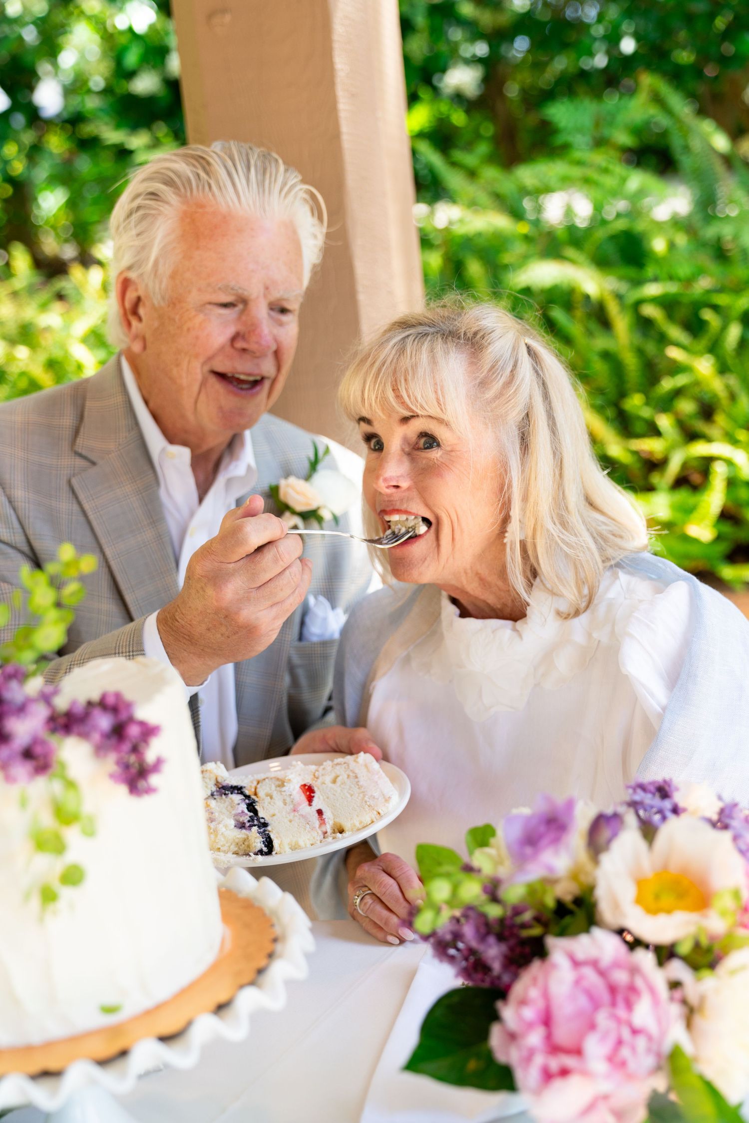 Senior couple enjoying wedding cake together outdoors with colorful floral arrangements on table.
