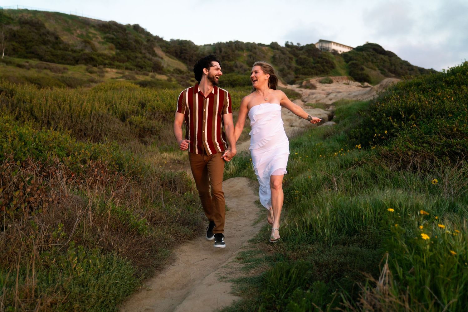 Two people in casual attire walk hand in hand along a scenic coastal trail surrounded by green vegetation at sunset.