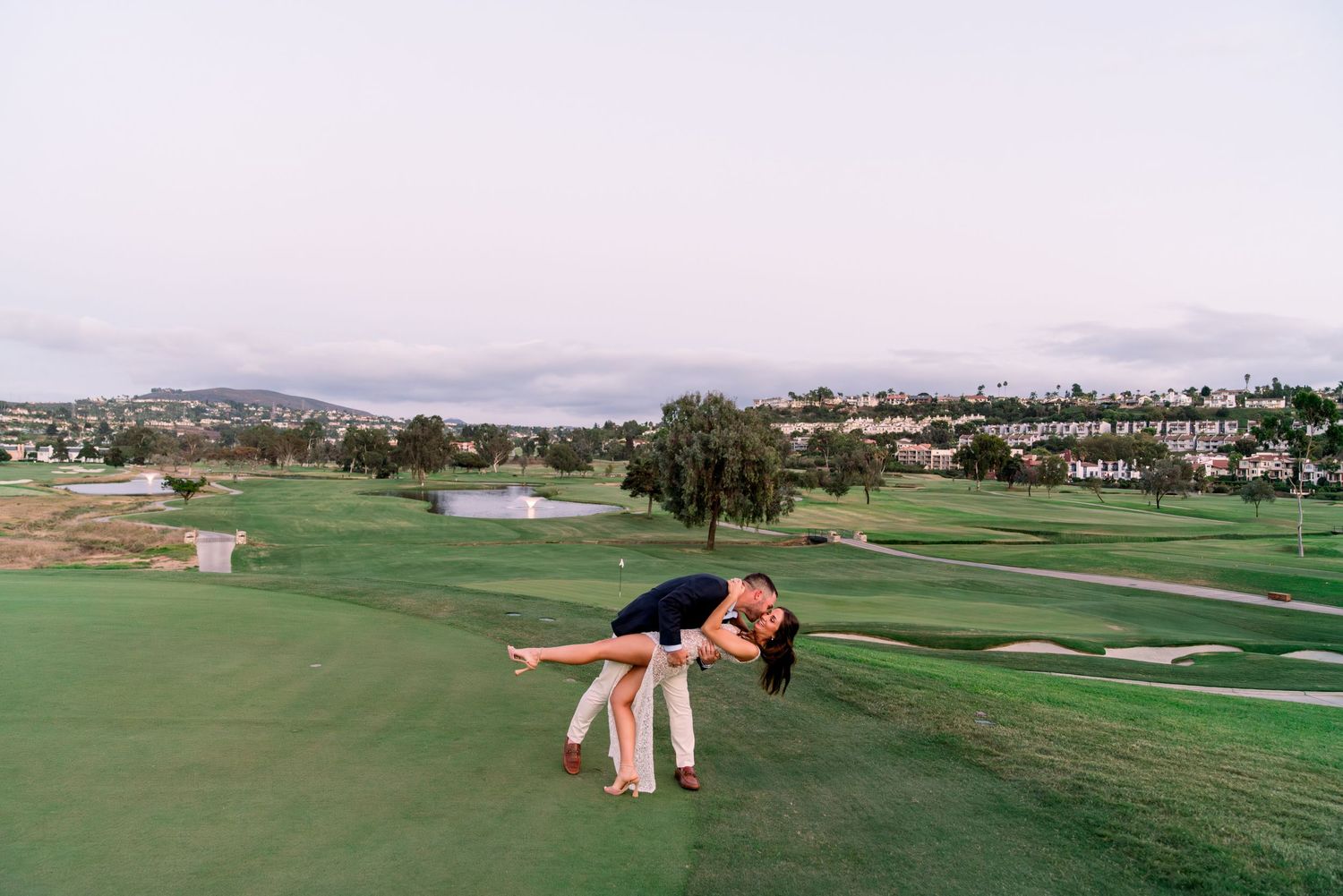 Playful couple striking a dramatic pose on a golf course with mountains in the background.