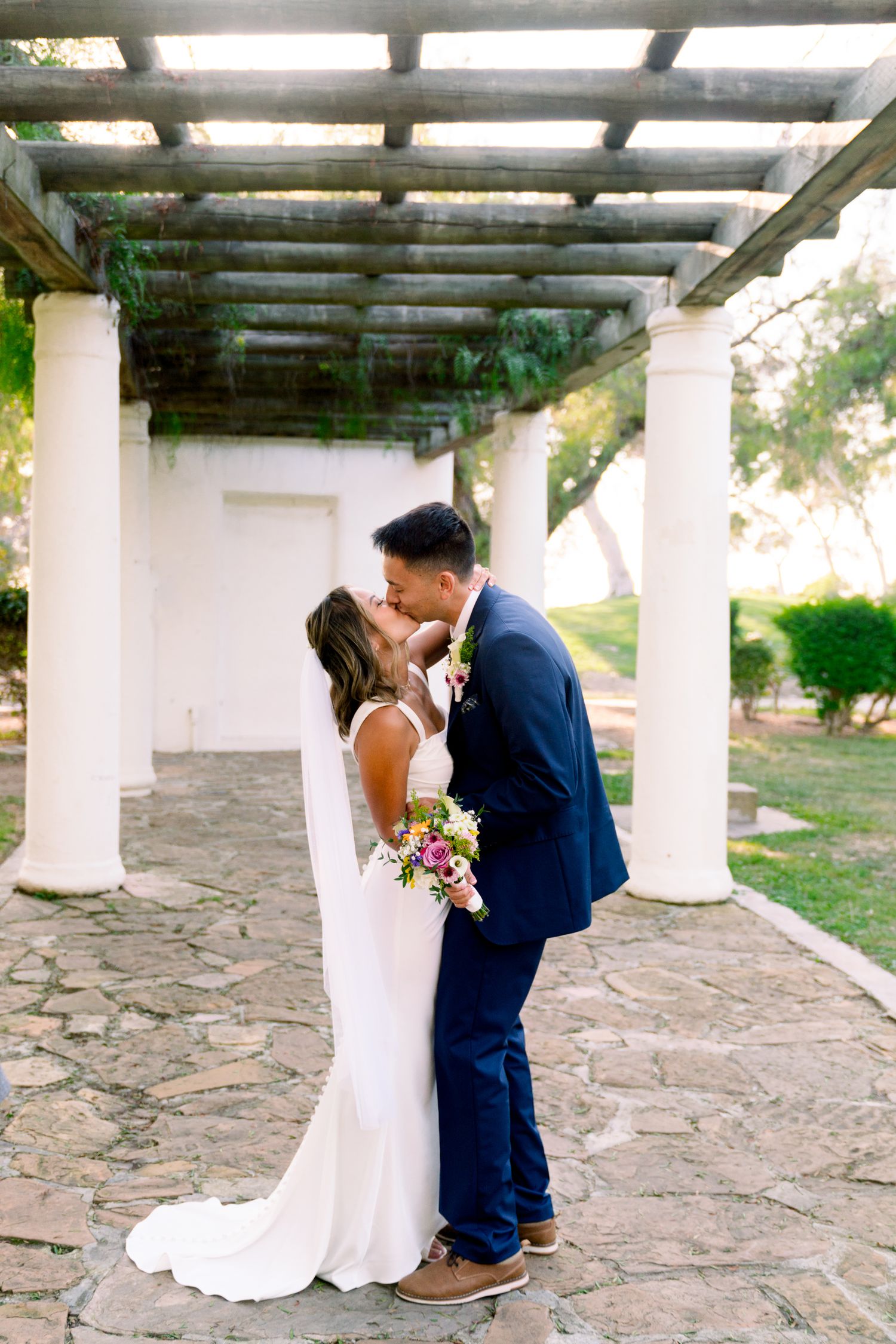 A romantic wedding couple shares a kiss under a white columned pergola on a stone patio.