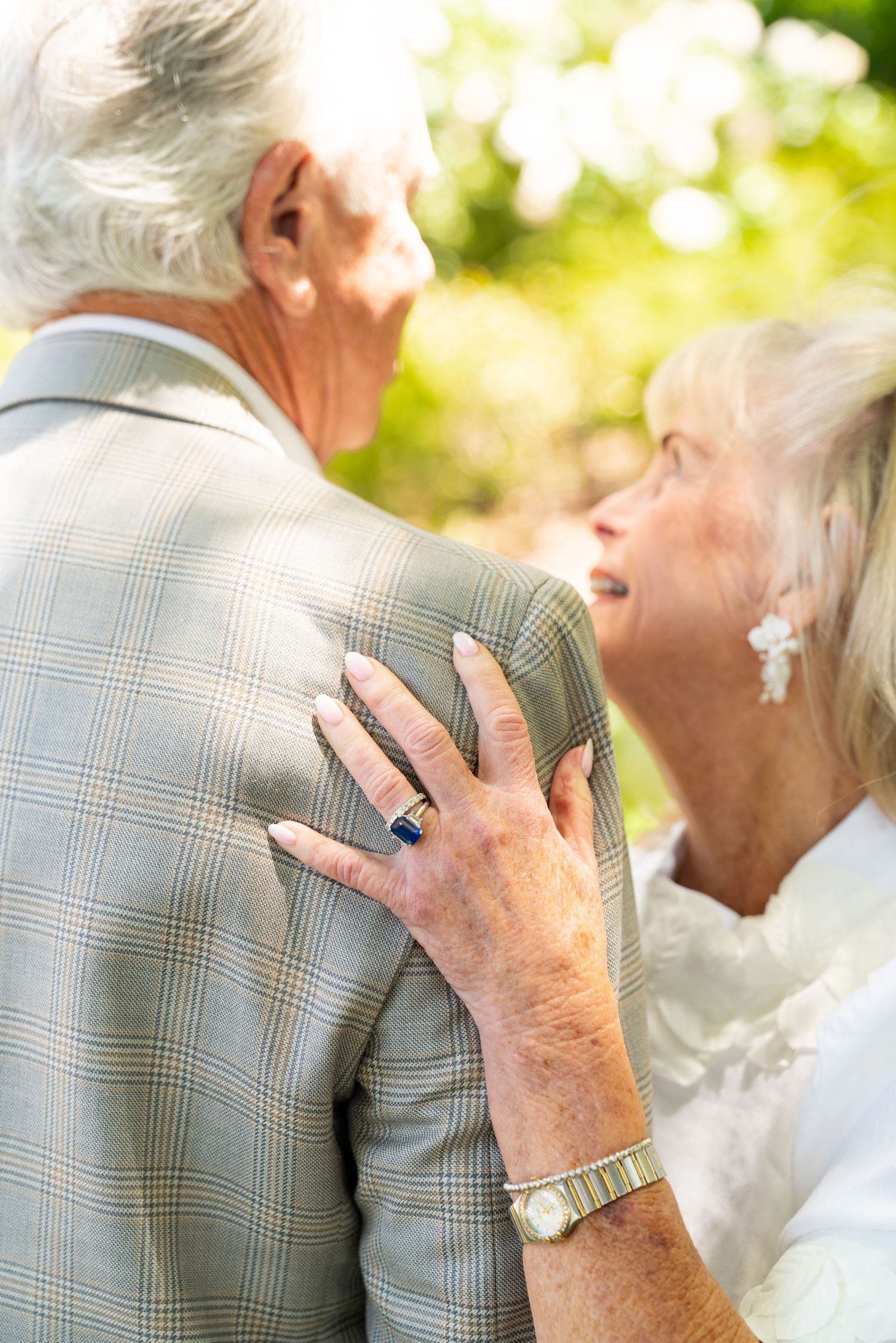 An elderly couple shares an intimate moment outdoors with the woman's hand resting on her partner's plaid jacket.