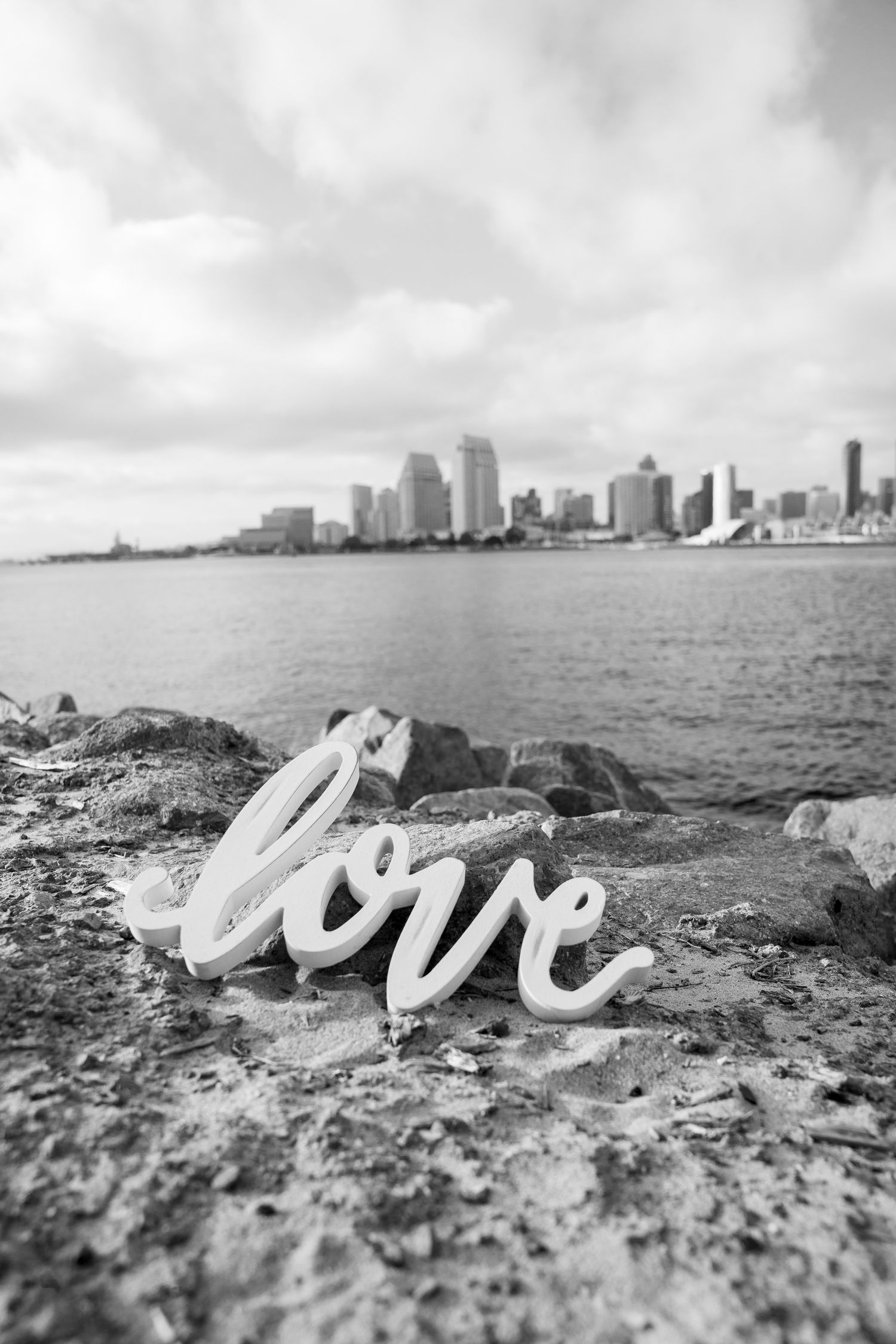 Black and white photo of 'love' text with city skyline across water in background.